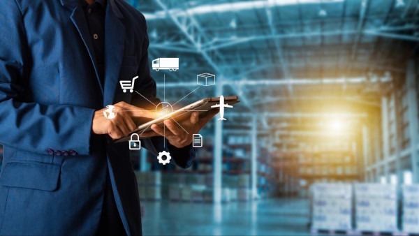 Man in a suit in a warehouse on a tablet with images of different supply chain processes displayed from his tablet