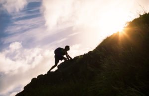 A person climbing on the side of a mountain with the sun glaring in the background