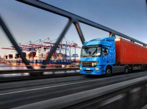 Truck hauling a cargo-container driving along a bridge near a port with a cargo ship in the background