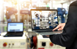 Man holding a laptop in a factory setting with the screen showing multiple graphs and metrics